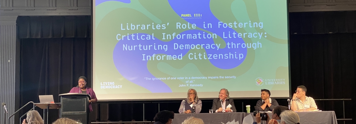 Photo of Twanna Hodge speaking at a podium in front of a projected slide titled "Libraries' Role in Fostering Critical Information Literacy: Nurturing Democracy Through Informed Citizenship." Three panelists, including Dr. Ana Ndumu, sit at a table before an audience.