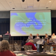 Photo of Twanna Hodge speaking at a podium in front of a projected slide titled "Libraries' Role in Fostering Critical Information Literacy: Nurturing Democracy Through Informed Citizenship." Three panelists, including Dr. Ana Ndumu, sit at a table before an audience.