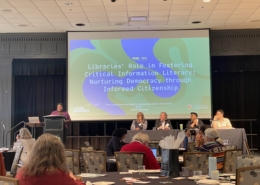 Photo of Twanna Hodge speaking at a podium in front of a projected slide titled "Libraries' Role in Fostering Critical Information Literacy: Nurturing Democracy Through Informed Citizenship." Three panelists, including Dr. Ana Ndumu, sit at a table before an audience.