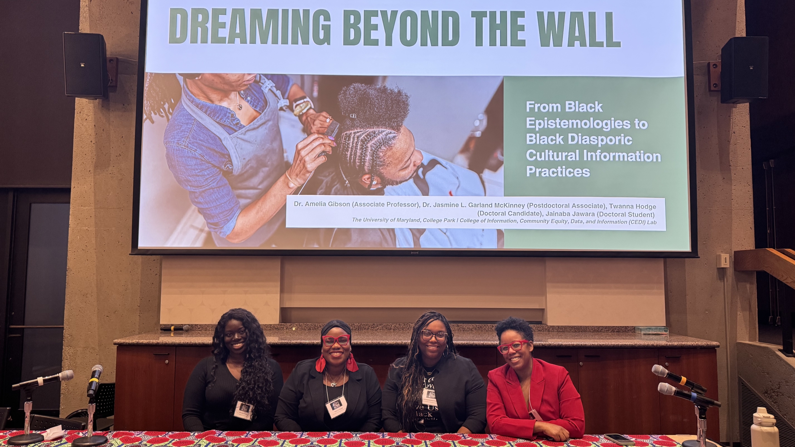 Four women-Drs. Amelia Gibson and Jasmine Garland McKinney, and PhD students Twanna Hodge, and Jainaba Jawara sit at a table in front of a powerpoint presentation. The screen reads, "Dreaming beyond the wall: From Black epistemologies to Black diasporic cultural information practices."
